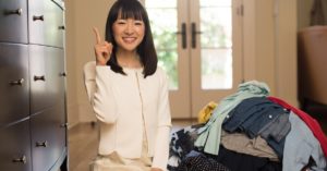 Marie Kondo stands next to a pile of clothes, smiling, with one finger pointing up